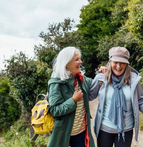 Zwei Freundinnen wandern lachend mit Rucksäcken auf sonnigem Waldweg