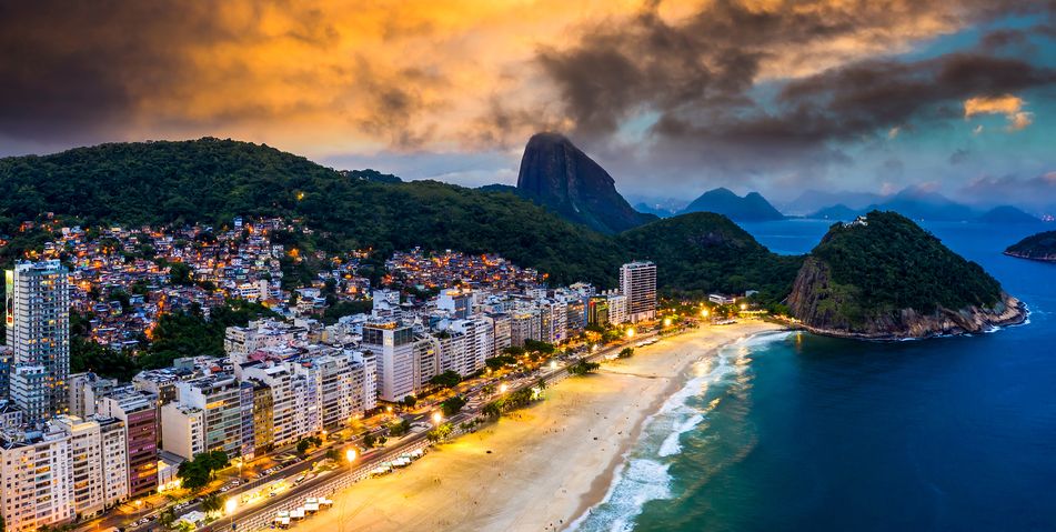Copacabana-Strand in Rio de Janeiro bei Sonnenuntergang mit beleuchteter Promenade und dem Zuckerhut im Hintergrund