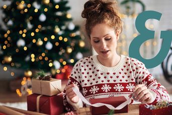 Eine Frau mit einem typischen Weihnachtspulli packt gemütlich Weihnachtsgeschenke ein mit dem Berge & Meer Superzeichen