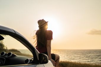 Frau sitzt auf der Motorhaube eines Autos und blickt auf den Sonnenuntergang am Meer