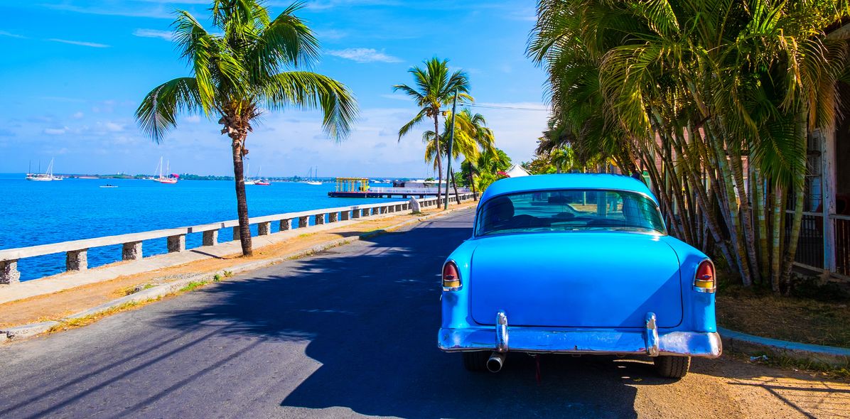 Blauer Oldtimer in Kuba auf einer Küstenstraße mit Palmen und Meer