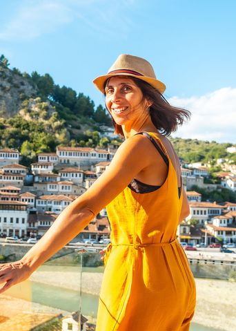 Frau in gelber Kleidung mit Hut steht an einem Geländer vor dem Stadtpanorama von Berat in Albanien