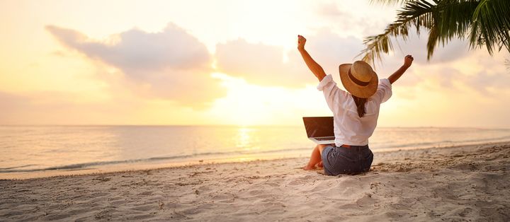 Frau sitzt mit Laptop am Strand vor dem Meer und genießt den Sonnenaufgang