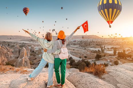 Zwei Reisende stehen in Kappadokien vor Felslandschaft und aufsteigenden Heißluftballons