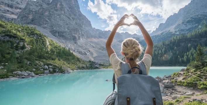 Frau von hinten fotografiert, steht vor einer weitläufigen Seenlandschaft in Italien und fängt mit ihrer zum Herz geformten Hand die Sonne ein