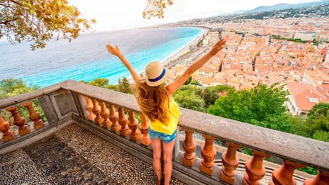 Frau mit Hut und ausgestreckten Armen blickt von einer Aussichtsterrasse über die Küste und Stadt von Nizza