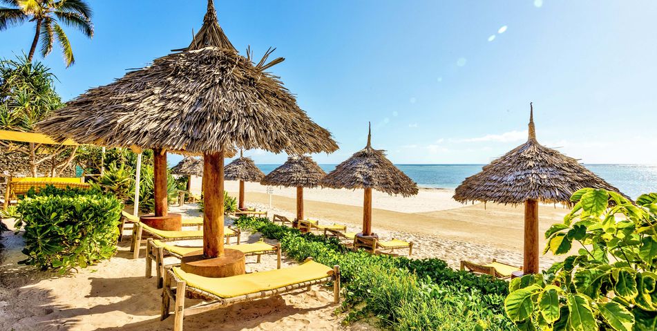 Blick aufs Meer von Sansibar über einen typischen Strand mit Sonnenschirmen und -liegen aus Palmen und Bäumen