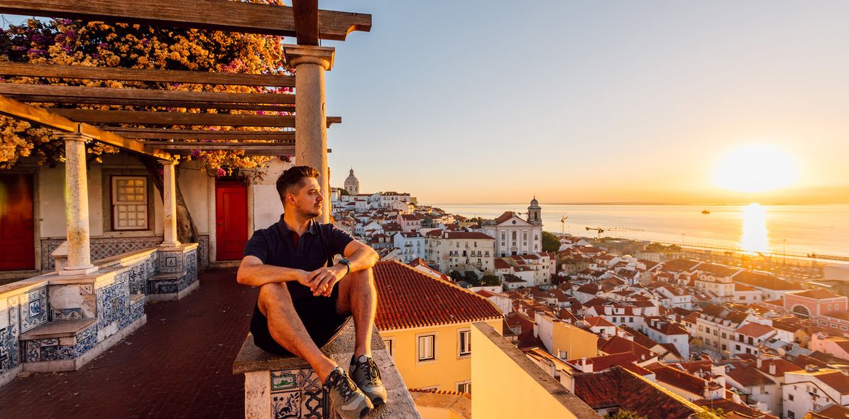 Ein Mann sitzt auf einer Terrasse über den Dächern von Lissabon und blickt auf den Sonnenuntergang