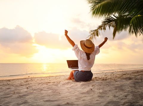 Frau sitzt mit Laptop am Strand vor dem Meer und genießt den Sonnenaufgang