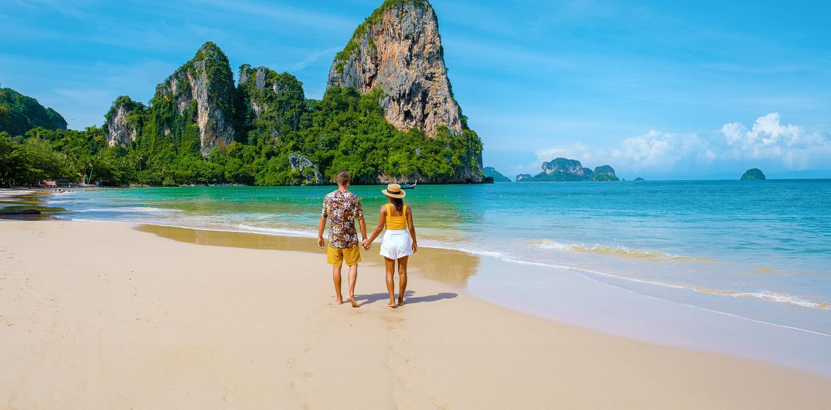 Paar händchenhaltend von hinten am Strand von Krabi in Thailand mit Blick auf das Meer und Felsen im Hintergrund 