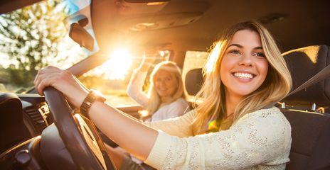Zwei Frauen sitzen in einem Auto und lächeln in die Kamera während die Sonne ins Auto scheint