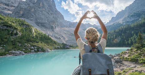Frau von hinten fotografiert, steht vor einer weitläufigen Seenlandschaft in Italien und fängt mit ihrer zum Herz geformten Hand die Sonne ein