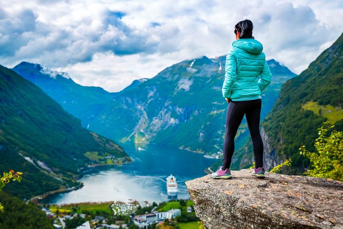 Eine Frau steht auf einem Felsen und schaut auf einen Fjord hinab, indem ein Kreuzfahrtschiff fährt