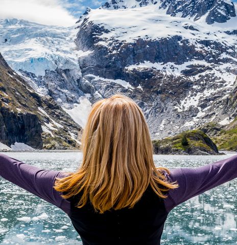 Reisende blickt mit ausgebreiteten Armen auf schneebedeckte Berge und einen Gletscher im Kenai-Fjords-Nationalpark in Alaska