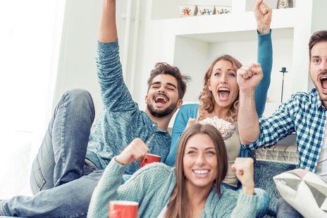 Eine Gruppe von jungen Menschen sitzt jubelnd mit Getränken und Snacks in einem Wohnzimmer und schaut vermutlich in einen Fernseher