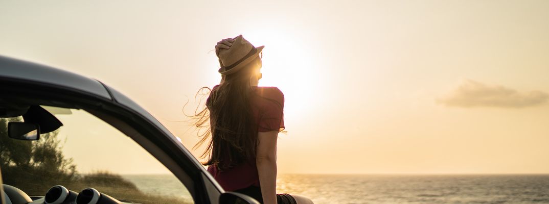 Frau sitzt auf der Motorhaube eines Autos und blickt auf den Sonnenuntergang am Meer