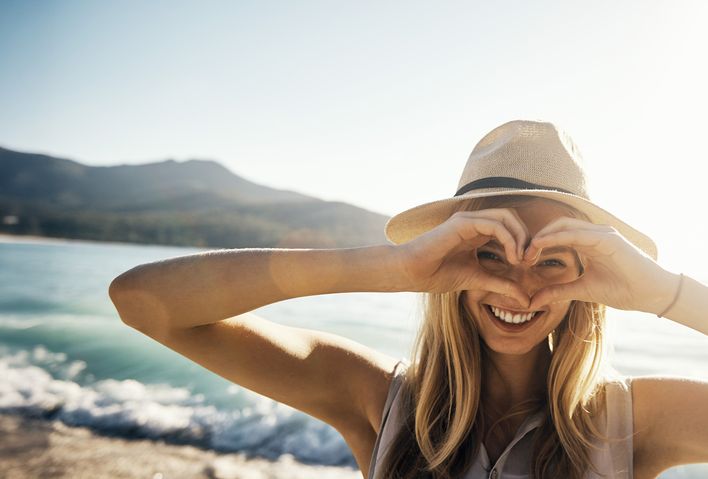 Frau mit Sommerhut am Strand formt mit den Händen ein Herz und schaut hindurch