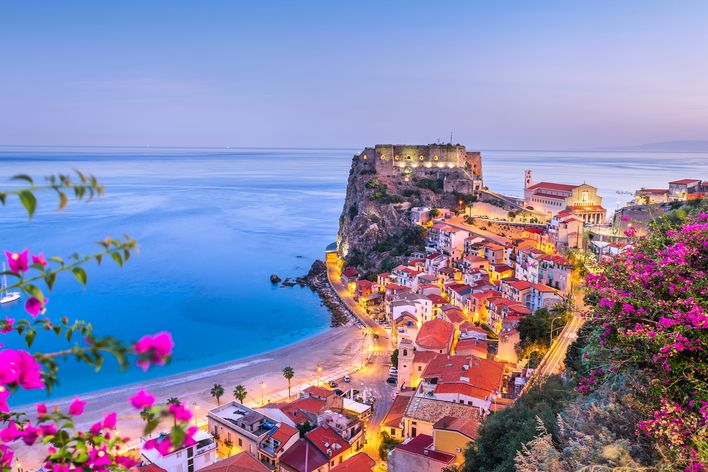 Panoramablick auf die Küste von Scilla in Kalabrien mit bunten Häusern am Hang, blauem Meer und pink blühenden Bougainvillea im Vordergrund