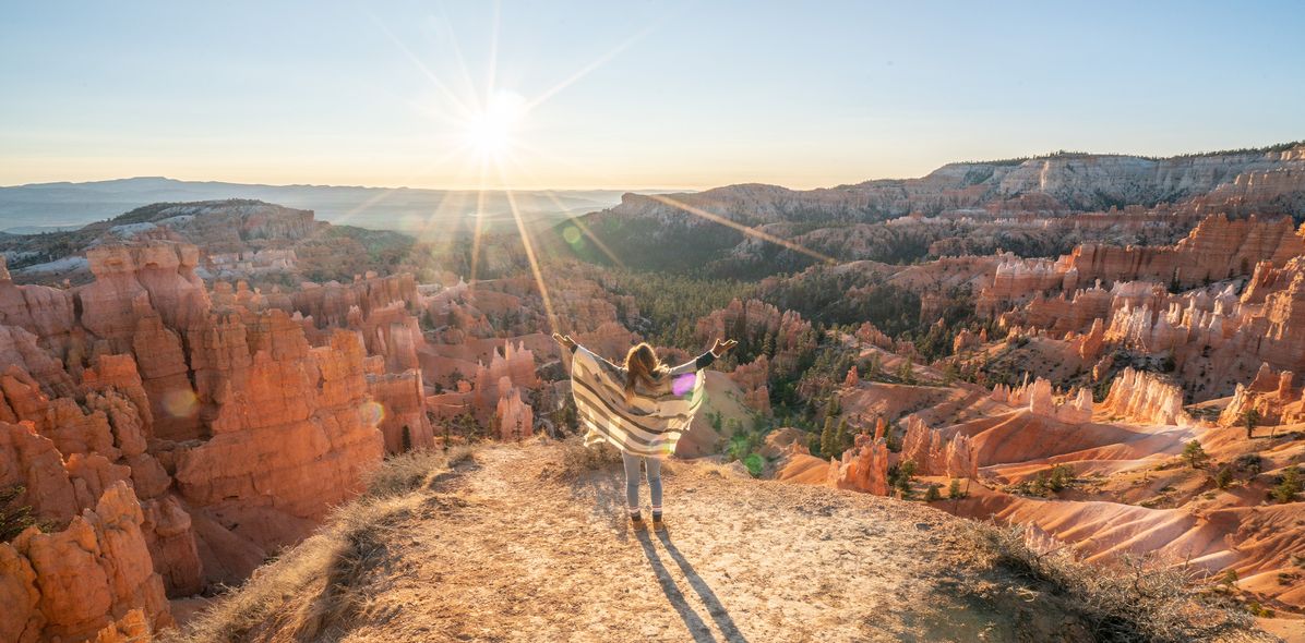 Frau in Rückansicht mit ausgebreiteten Armen blickt über den Bryce Canyon in den USA in den Sonnenuntergang