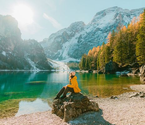 Frau sitzt auf einem Felsen vor einem See umgeben von sonnenbestrahlten Bergen und Bäumen in Südtirol