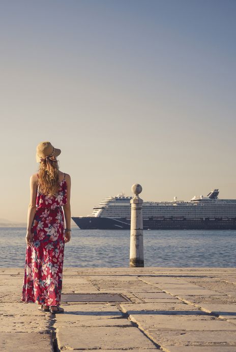 Frau mit einem bunten langen Kleid steht an einer Promenade und schaut auf ein Kreuzfahrtschiff