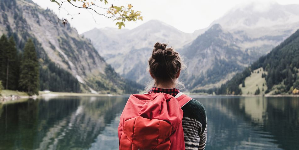 Blick auf die Rückansicht einer Frau mit Rucksack, die vor einem Bergsee auf die Berge blickt