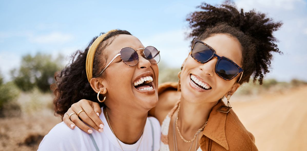 Zwei Freundinnen lachen gemeinsam in der Sonne und tragen Sonnenbrillen