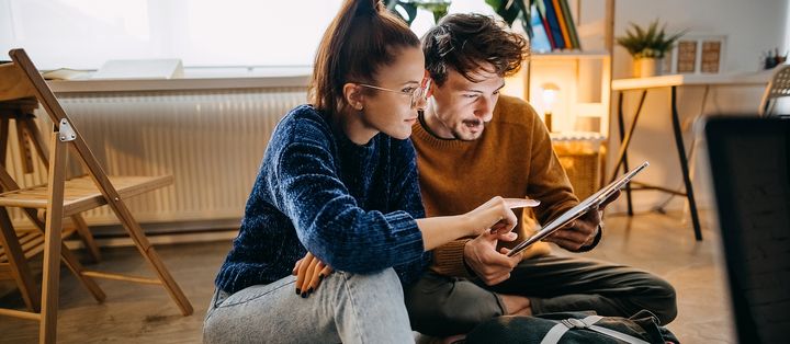 Paar sitzt auf dem Boden im Wohnzimmer und schaut interessiert auf ein Tablet