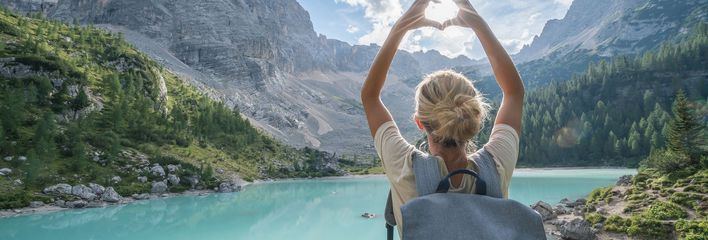 Frau von hinten fotografiert, steht vor einer weitläufigen Seenlandschaft in Italien und fängt mit ihrer zum Herz geformten Hand die Sonne ein