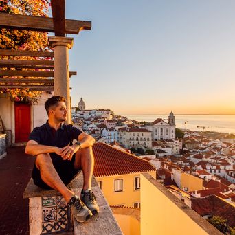 Ein Mann sitzt auf einer Terrasse über den Dächern von Lissabon und blickt auf den Sonnenuntergang