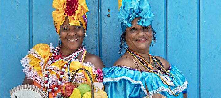 Kubanische Frauen mit Obstkorb und Fächer in traditioneller Kleidung vor blauer Holzwand