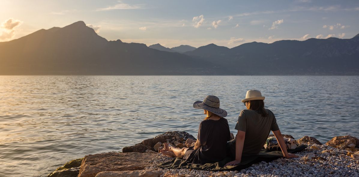 Paar sitzt am Ufer des Gardasees und genießt den Sonnenuntergang mit Blick auf die Berge