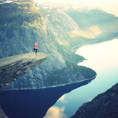 Eine Person steht auf der Trolltunga und im Hintergrund sind Berge, Fjorde und Gletscher zu sehen