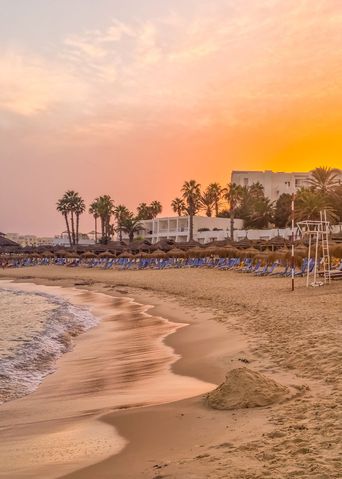 Goldener Sandstrand mit strohbedeckten Sonnenschirmen umringten Liegen vor der Brandung bei Abendlicht in Monastir in Tunesien