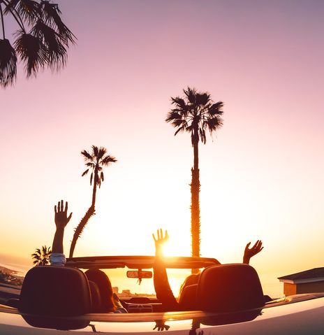 Zwei Frauen sitzen in einem Cabrio welches der Sonne entgegen und von Palmen umgeben auf einem Parkplatz steht und die Hände in die Höhe strecken