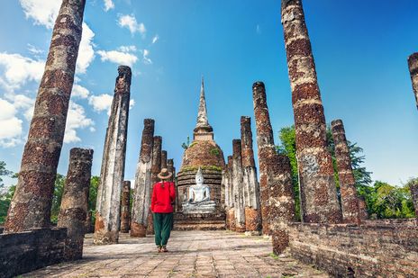 Touristin mit roter Kleidung und Hut steht vor dem Sukhothai Tempel in Thailand