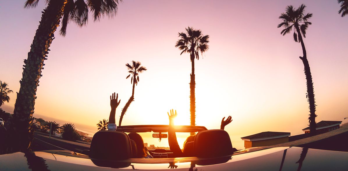 Zwei Frauen sitzen in einem Cabrio welches der Sonne entgegen und von Palmen umgeben auf einem Parkplatz steht und die Hände in die Höhe strecken
