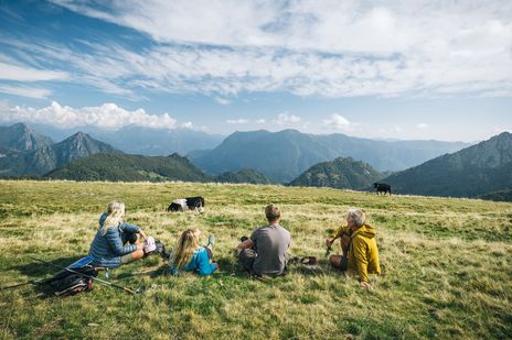 Wandergruppe genießt Rast auf grüner Almwiese mit Bergpanorama im Hintergrund