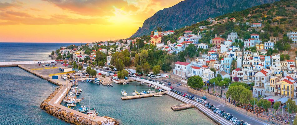 kleiner Hafenort auf einer griechischen Insel mit bunten Häusern am Hang und Sonnenuntergang im Hintergrund