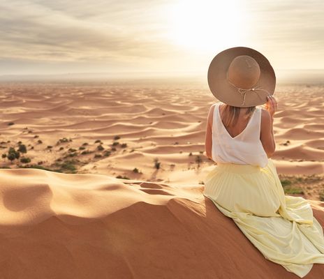 Frau mit Sonnenhut sitzt in der Sahara in Marokko und blickt auf die weite Landschaft