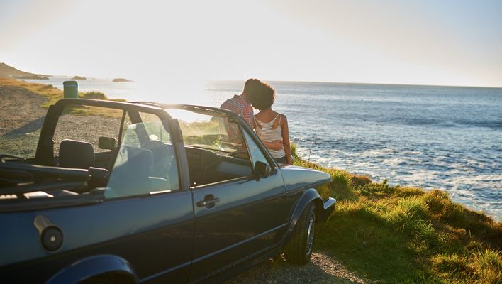 Paar lehnt an der Motorhaube ihres Cabrios und blickt auf das Meer
