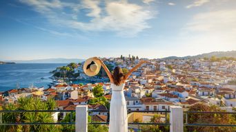 Eine Frau im weißen Kleid mit einem Hut in der Hand steht auf einer Aussichtsplattform und schaut in die Stadt Skiathos in Griechenland