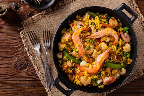 Blick von oben auf die typisch spanische Speise Paella in einer gusseisernen Pfanne auf einem Tischset, umrahmt von Gabeln und einer Pfeffermühle