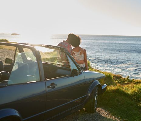 Paar lehnt an der Motorhaube ihres Cabrios und blickt auf das Meer