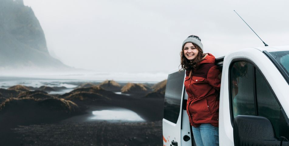 Frau steht laechelnd vor Mietwagen in einer nebligen Landschaft in Island