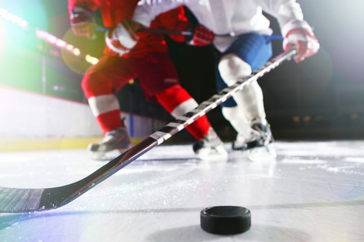 Die Beine von zwei Eishockeyspielern hinter einem der Schläger, der kurz davor ist, den Puck zu treffen