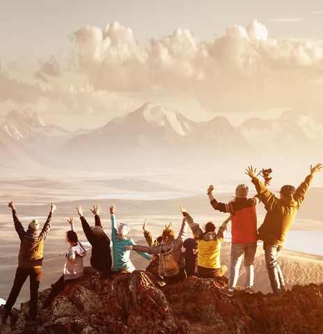 Gruppe von Wanderern steht auf einem Felsen vor einer Berglandschaft und reißt jubelnd die Arme nach oben