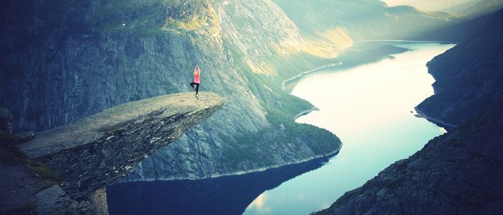 Eine Person steht auf der Trolltunga und im Hintergrund sind Berge, Fjorde und Gletscher zu sehen