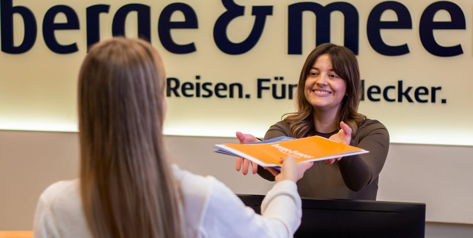 Eine Person überreicht einer Kollegin Ihre Unterlagen am Empfang von Berge & Meer