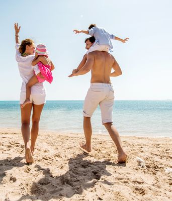 Paar mit zwei Kinder in den Armen läuft am Strand lachend Richtung Meer